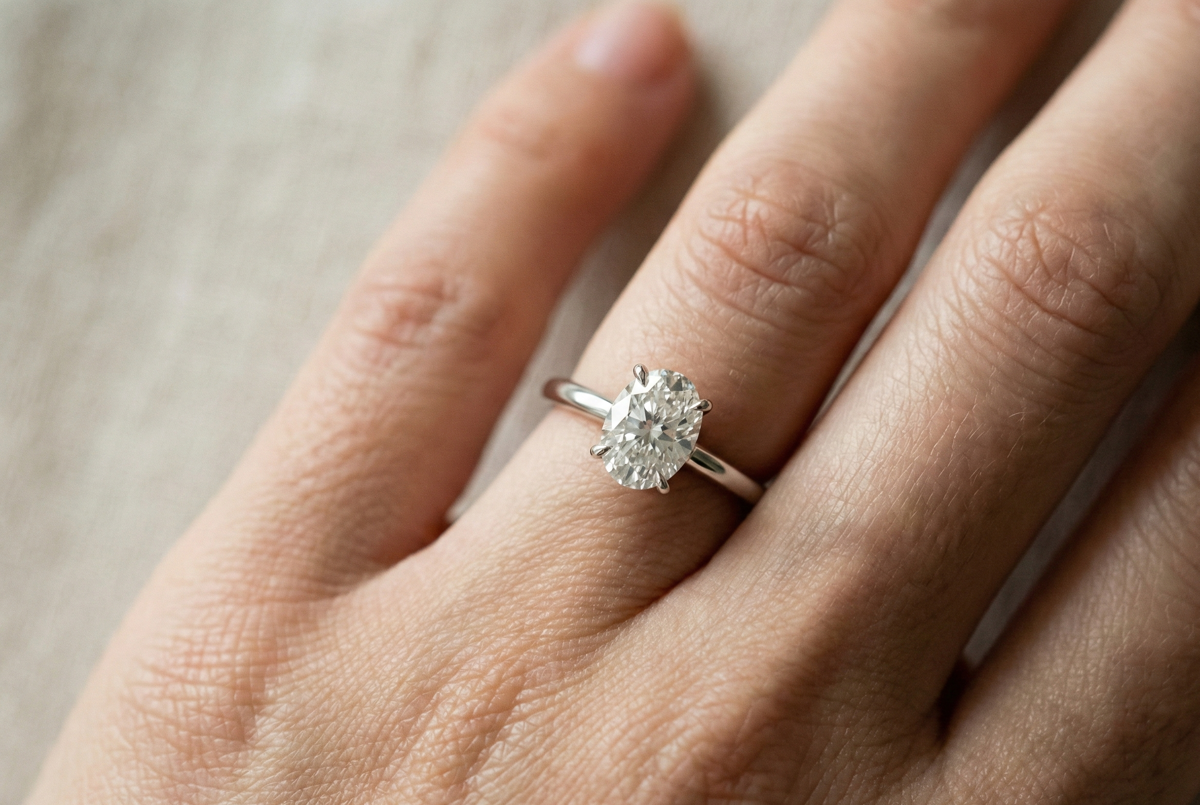 Close-up of a woman’s hand wearing a minimalist bespoke engagement ring featuring a sparkling oval-cut diamond on a slim 1.7mm tapered band, captured in soft natural lighting with a high-end editorial aesthetic.
