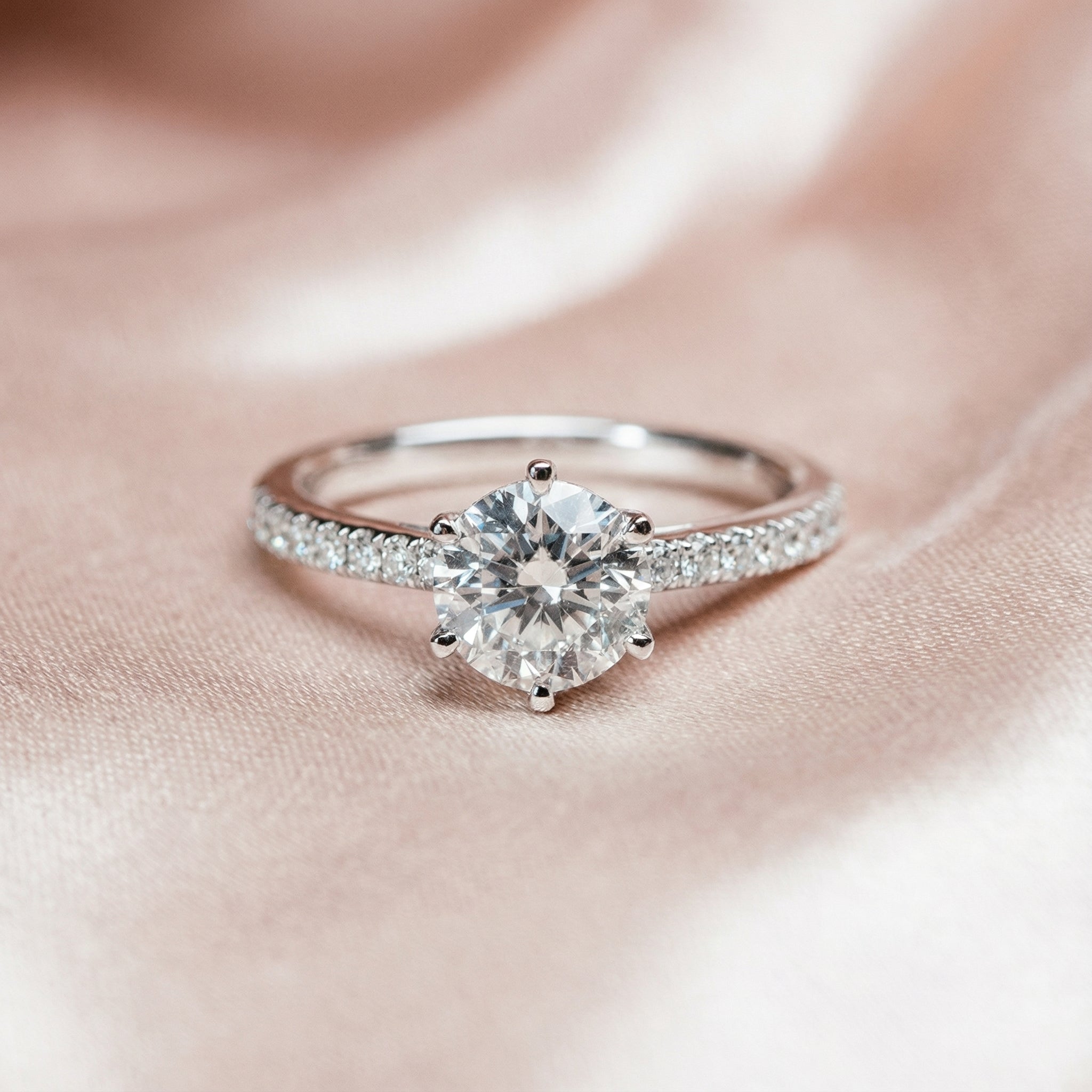 Round diamond engagement ring with pavé band on soft blush fabric background.