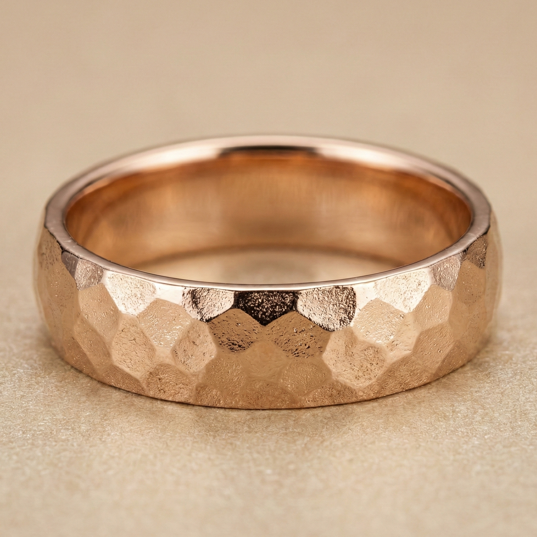 18K rose gold men’s hammered wedding band featuring a rugged faceted texture and high-polish interior, photographed in warm neutral studio lighting.