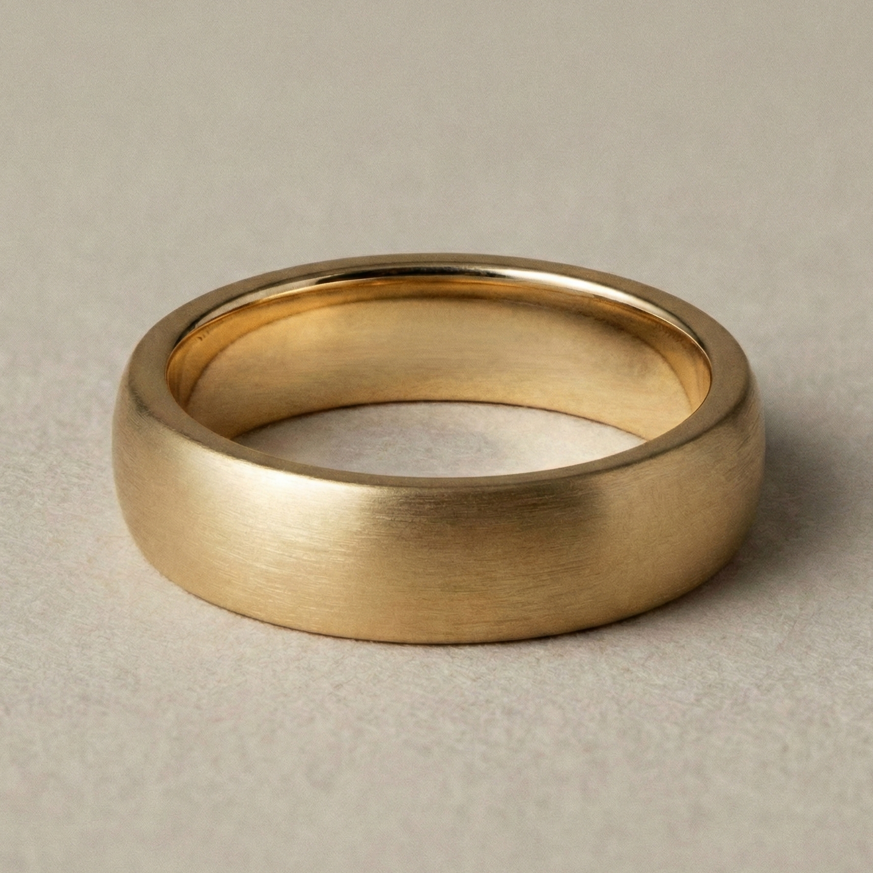 18K yellow gold men’s wedding band with a soft matte brushed finish and rounded comfort-fit interior, photographed in warm neutral studio lighting.