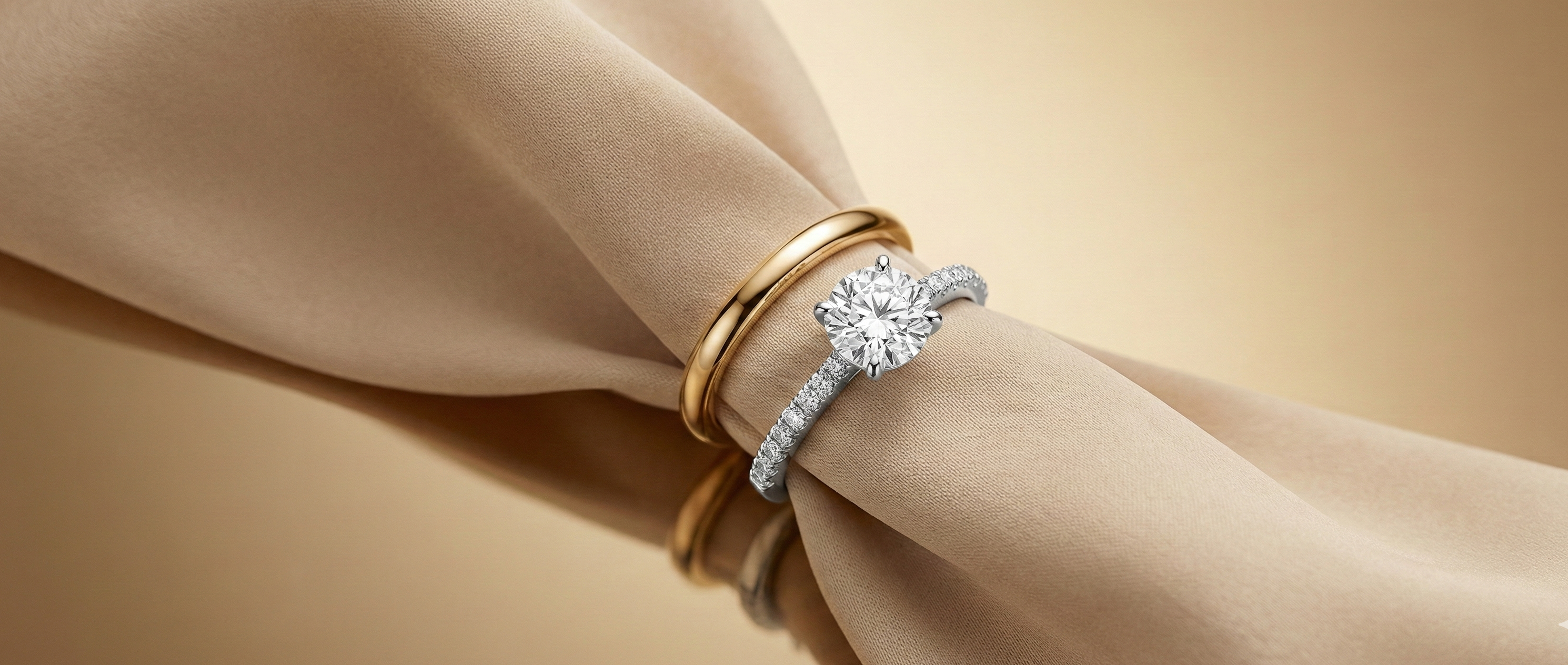 Solitaire engagement ring with a diamond pavé band paired with 18K yellow gold wedding bands, styled on soft beige fabric for a luxurious studio look
