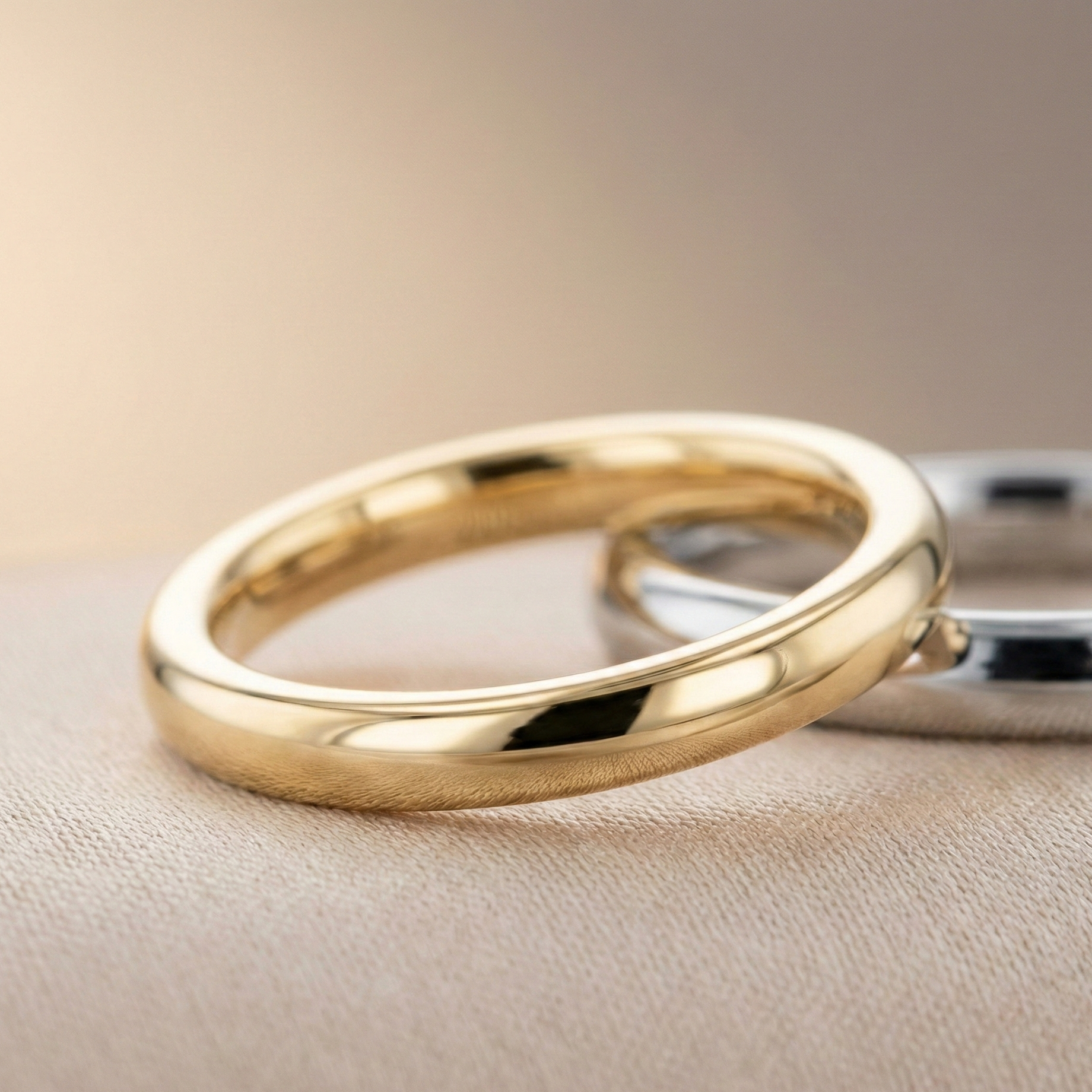 18K yellow gold classic wedding band with a high-polish rounded profile, photographed in soft beige studio lighting with a platinum band in the background.