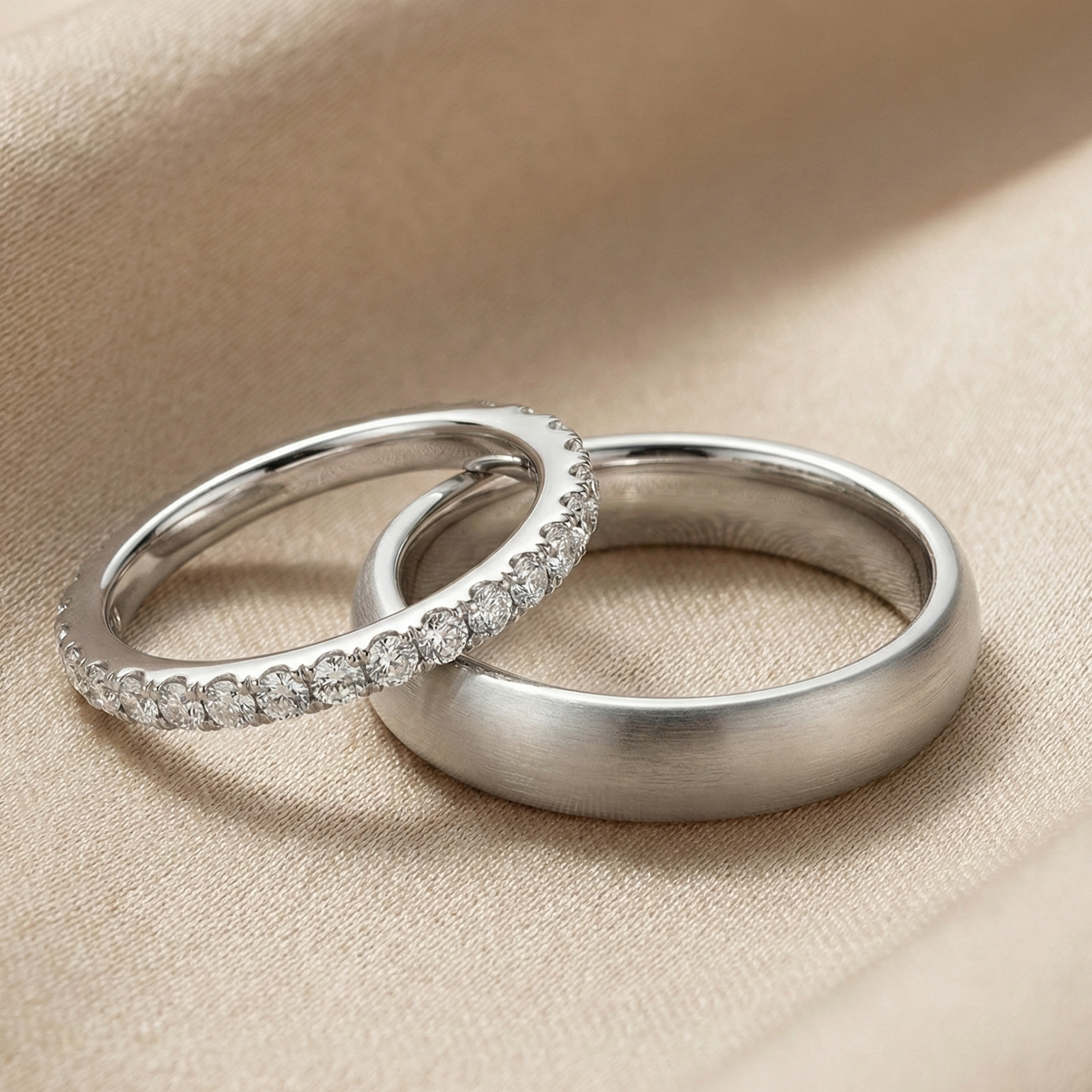 Platinum couple wedding bands featuring a slim pavé diamond band stacked with a wide matte-finish men’s band, photographed in soft champagne studio lighting on a fabric backdrop.